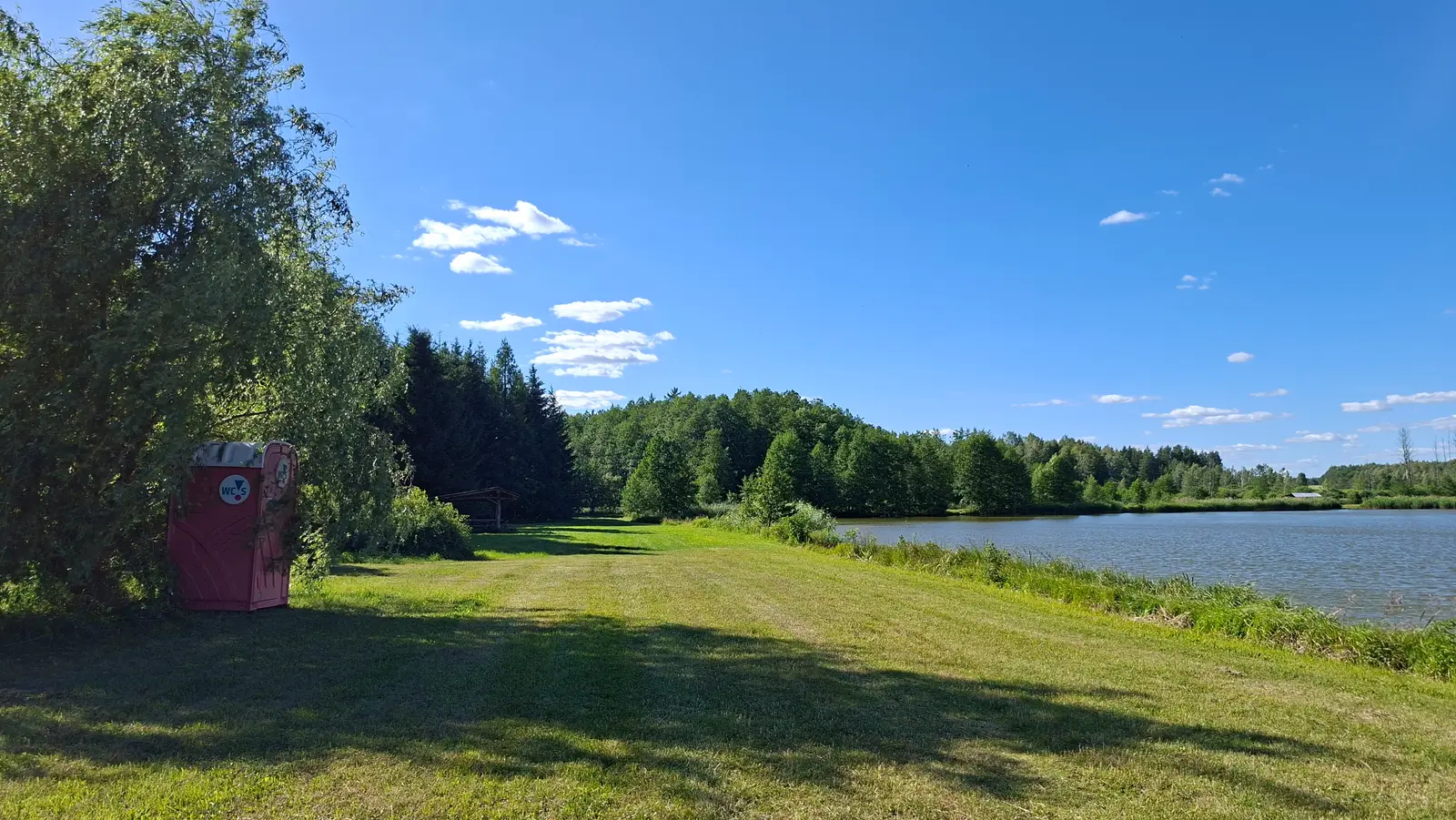 Zdjęcie przedstawia sielski krajobraz w orientacji poziomej, gdzie na pierwszym planie po lewej stronie stoi czerwona przenośna toaleta typu toi-toi, umieszczona w cieniu dużego, rozłożystego drzewa. Przed obiektem rozpościera się rozległy, zielony trawnik, który prowadzi w stronę spokojnego jeziora znajdującego się po prawej stronie kadru. W tle widoczna jest gęsta linia lasu iglastego i liściastego, a całość dopełnia intensywnie niebieskie, bezchmurne niebo z delikatnymi obłokami. Kompozycja zdjęcia opiera się na kontraście między słoneczną murawą a zacienionymi fragmentami przy drzewie, co nadaje zdjęciu głębi. Na samej toalecie widoczne jest wyraźne logo „WC S”, stanowiące jedyny akcent tekstowy w tej przyrodniczej scenerii.